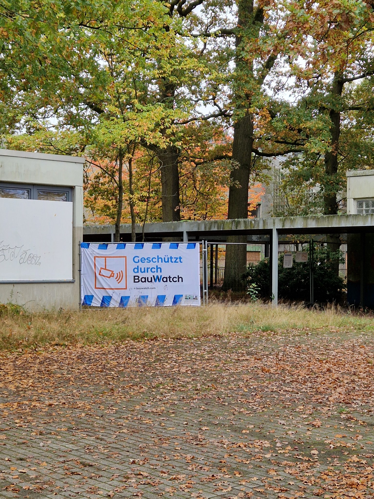 BauWatch-Überwachungsbanner an der ehemaligen Gerhart-Hauptmann-Realschule, angebracht an einer Außenwand im herbstlich bewachsenen Schulhof.