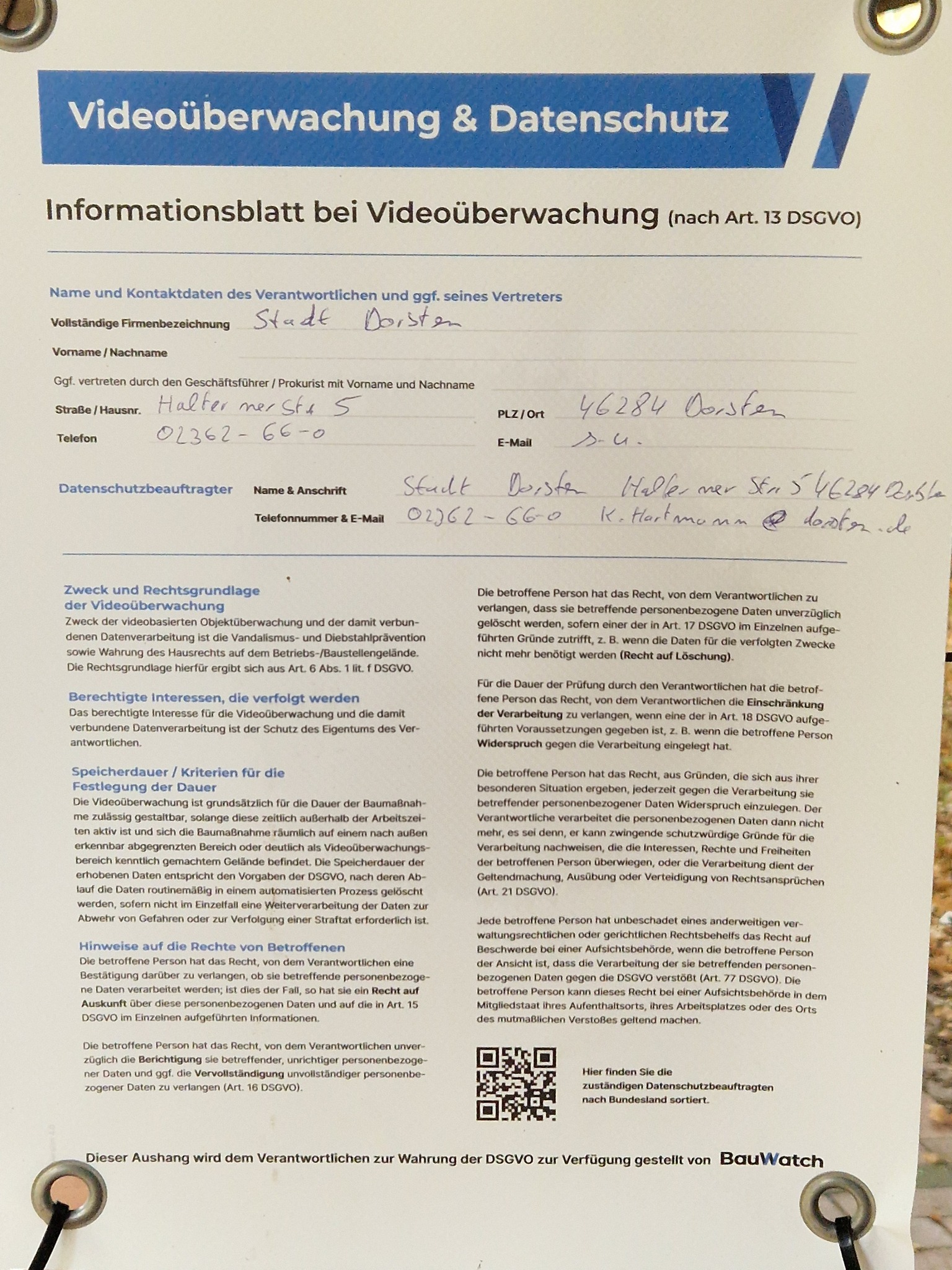 Mit Graffiti besprühtes BauWatch-Sicherheitsbanner am verschlossenen Eingangstor des Schulgeländes, umgeben von Herbstlaub und Bäumen.